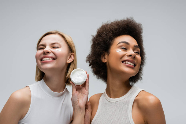 pleased interracial models in white tops posing near jar of face cream isolated on grey - Foto, afbeelding