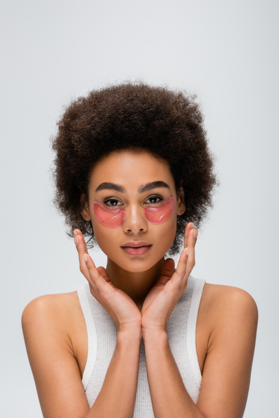 young african american woman in collagen eye patches holding hands near face while looking at camera isolated on grey - Photo, Image