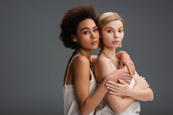 multiethnic blonde and brunette models with colorful visage looking at camera isolated on grey - Zdjęcie, obraz