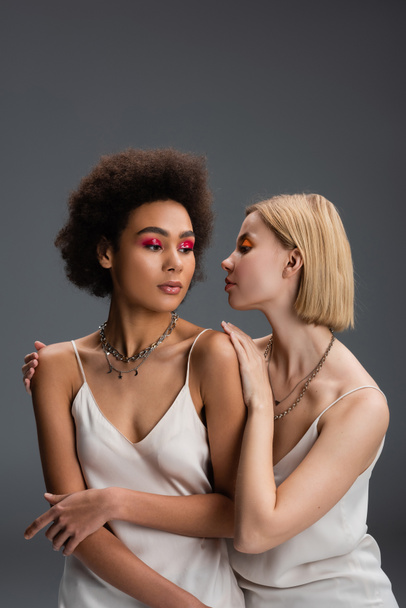 blonde woman touching shoulders of african american model in white strap dress and silver necklaces isolated on grey - Foto, imagen