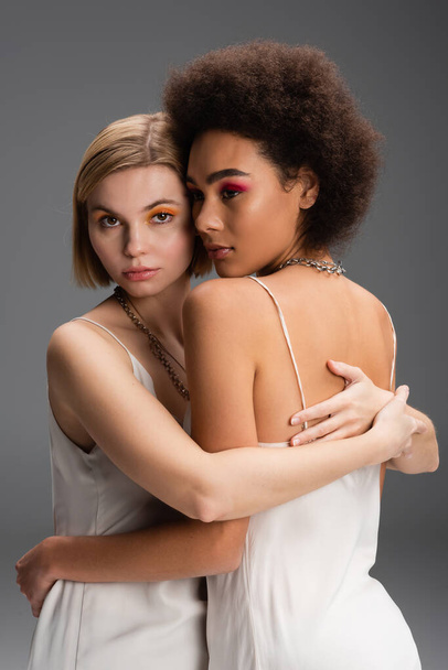 sensual interracial women in white strap dresses and colorful makeup embracing isolated on grey - Photo, Image