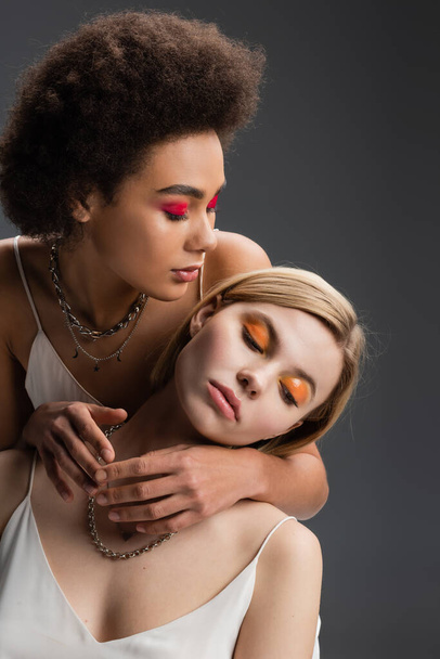 young african model in bright makeup and silver necklaces embracing blonde woman isolated on grey - Zdjęcie, obraz