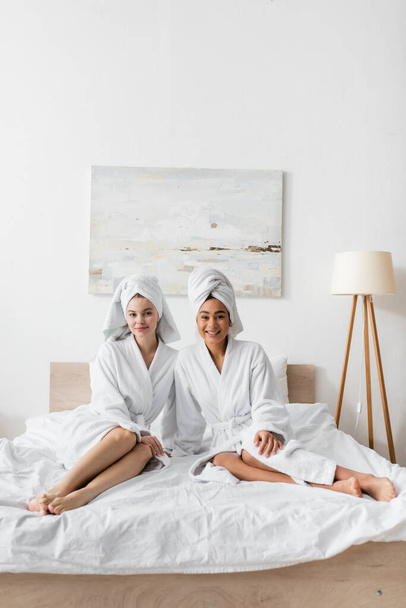 full length of young barefoot women in white soft robes and towels sitting on bed and smiling at camera - Foto, immagini
