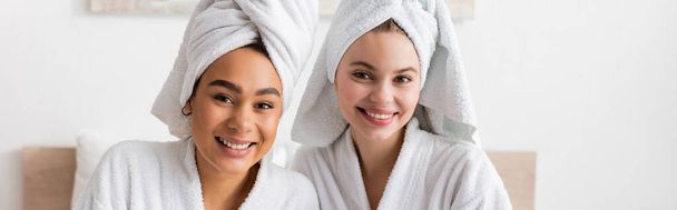jóvenes y positivas mujeres multiétnicas en suaves batas de felpa y toallas mirando a la cámara en el dormitorio, pancarta - Foto, imagen