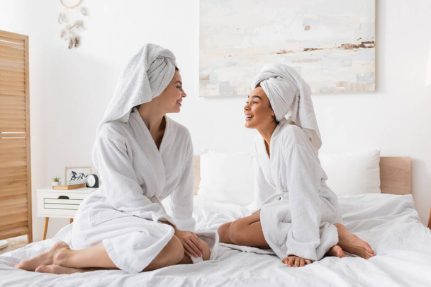 full length of barefoot interracial women in white robes and towels sitting on bed and smiling at each other - Foto, Imagem