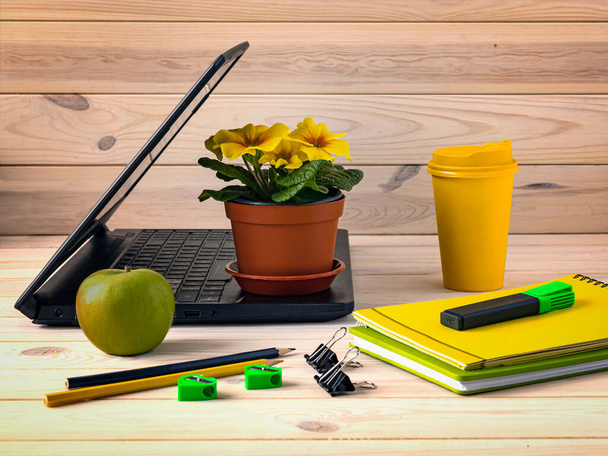 Kantoorruimte met open laptop, notitieblok, geel papieren kopje koffie, stift op houten tafel. Een groene appel en een pot met een bloem als decor. Bedrijfsachtergrond. - Foto, afbeelding