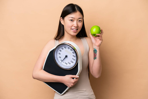 Young Chinese woman isolated on beige background with weighing machine and with an apple - Fotó, kép
