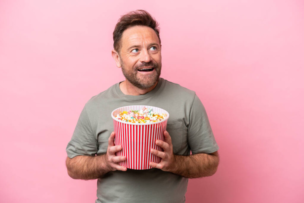 Middle age caucasian man isolated on pink background holding a big bucket of popcorns - Foto, afbeelding
