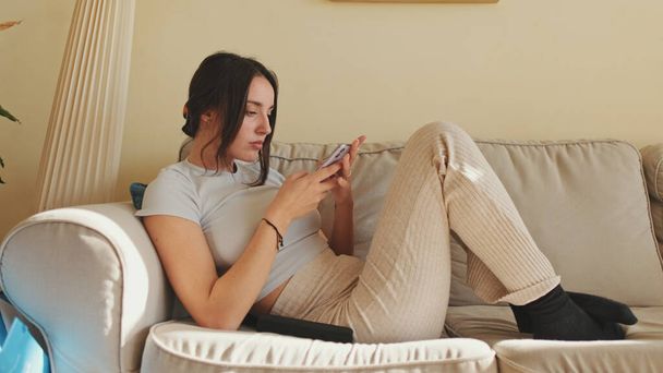 Young woman using mobile phone and changing channels from remote control while sitting on sofa at home - Фото, изображение