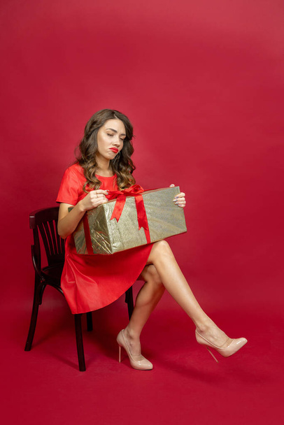 Donna bruna seduta sulla sedia delusa dal regalo in abito rosso su uno sfondo rosso - Foto, immagini