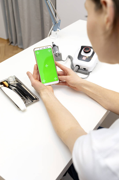 Manicure, pedicure technicus houdt gadget met groene chroma key screen, zittend op het werkstation in nagel schoonheid behandeling salon. Professioneel gereedschap staat op witte tafel. Studie manicure, verticaal - Foto, afbeelding