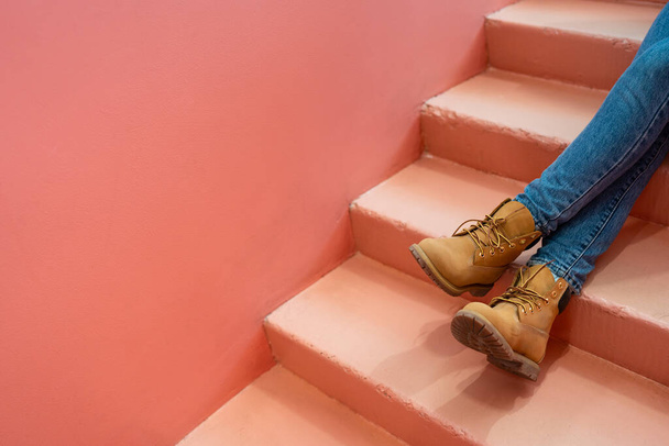 Ноги в синих джинсах и светло-коричневых сапогах вытянуты на кораллово-розовой лестнице к розово-оранжевой стене. Копировать пространство для рекламы, вставить текст или слоган. Расслабьтесь комфорт дизайн концепции цвета. - Фото, изображение