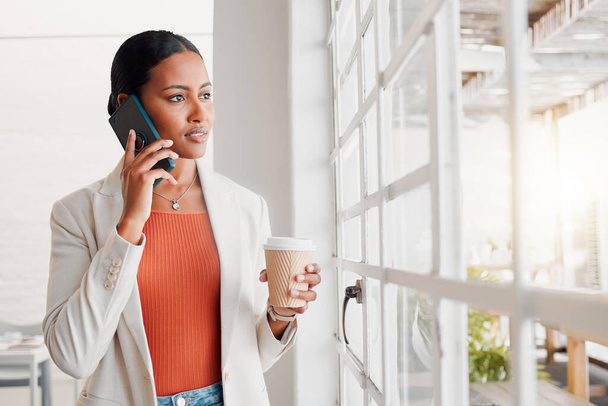 Young mixed race businesswoman on a call using her phone while drinking a coffee alone in an office at work. Confident hispanic businessperson looking out of a window drinking a coffee and talking on. - Photo, Image