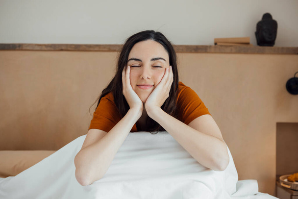 Dreamy attractive caucasian young woman sitting on comfortable bed in morning, smiling with closed eyes, resting chin on her hands. horizontal - Foto, afbeelding