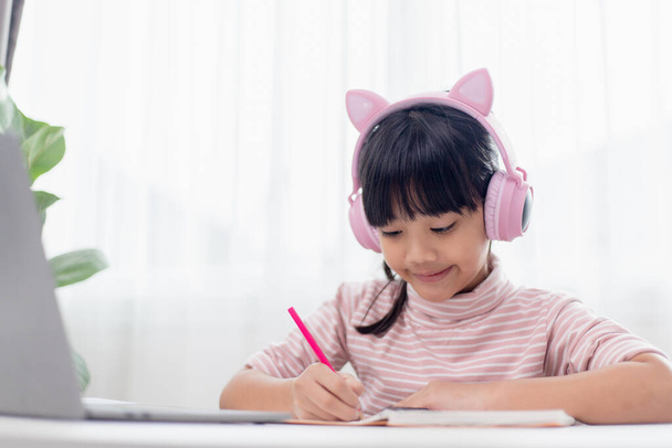 Aasialainen pieni tyttö kuulokkeissa on videopuhelu kaukainen luokka opettajan kanssa kannettavan tietokoneen avulla, opiskele verkossa tietokoneella, kotiopetuksen käsite - Valokuva, kuva