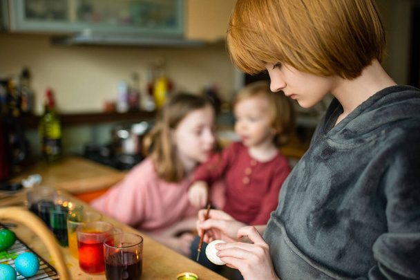 Two big sisters and their toddler brother dyeing Easter eggs at home. Children painting colorful eggs for Easter hunt. Kids getting ready for Easter celebration. Family traditions. - 写真・画像