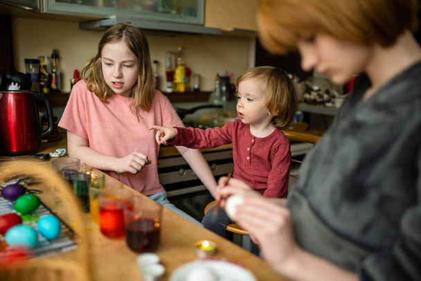 Two big sisters and their toddler brother dyeing Easter eggs at home. Children painting colorful eggs for Easter hunt. Kids getting ready for Easter celebration. Family traditions. - Фото, зображення