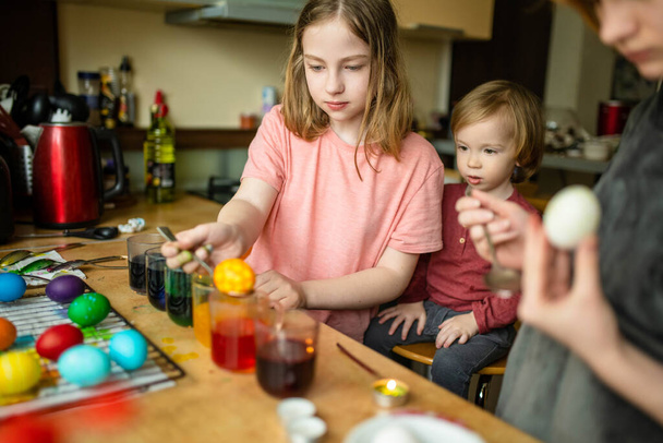 Two big sisters and their toddler brother dyeing Easter eggs at home. Children painting colorful eggs for Easter hunt. Kids getting ready for Easter celebration. Family traditions. - Photo, image