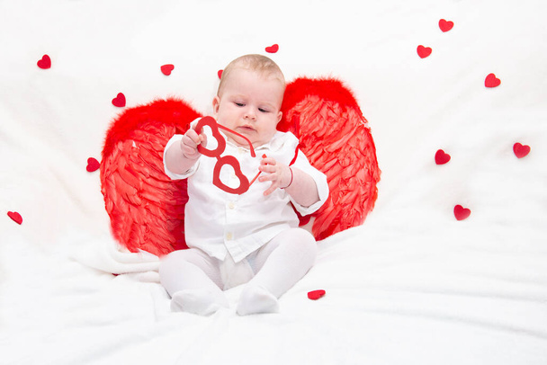 Un bebé con las mejillas hinchadas en ropa blanca con alas de cupido rojo, juega con gafas con corazones, aislado en una manta tierna blanca con corazones. cupid kid para un proyecto con amantes. - Foto, imagen