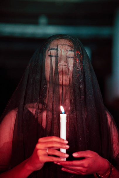 uma mulher asiática com os olhos fechados e um véu transparente manchado com sangue segura uma vela em sua mão durante a adoração satânica à noite - Foto, Imagem