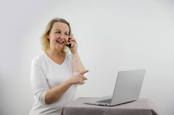woman shares on the phone news that she saw on the Internet. She points with a finger at a laptop on a white background, smiles, talks with friends, telling interesting facts about the story - Fotografie, Obrázek