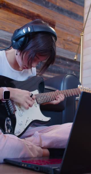 ノートパソコンのモニターの前でヘッドフォンでギターを持つ10代の少女の60 fpsの垂直4k映像。彼女は録音されたギターの音を聞いて. - 映像、動画