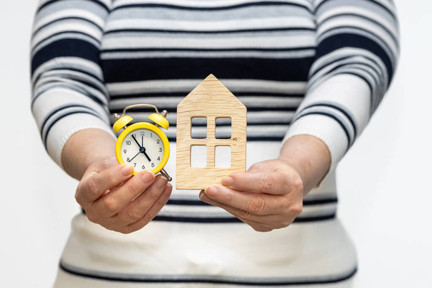 Γυναίκα κατέχει ξύλινη μινιατούρα σπίτι και ρολόι στα χέρια της, έννοια του χρόνου που απαιτείται για την επένδυση, αύξηση των τιμών υπό κατασκευή - Φωτογραφία, εικόνα