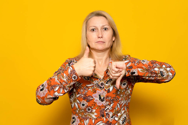 Pretty caucasian blonde woman in a patterned dress giving one thumbs up and one thumbs down isolated over yellow background. doubt concept - Photo, Image