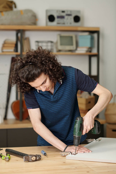 Young serious guy in blue shirt bending over table while drilling metallic hinge of plywood door of kitchen cabinet in garage of his house - Fotografie, Obrázek