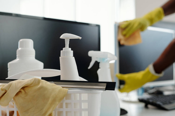 Group of detergents in white plastic bottles with dispensers and duster in basket against computer screen and gloved hands of man - Foto, Bild
