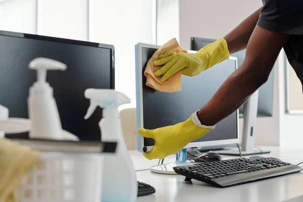 Mani guantate di giovane uomo afroamericano con spolverino che pulisce lo schermo del computer mentre si trova sul posto di lavoro del manager dell'ufficio - Foto, immagini