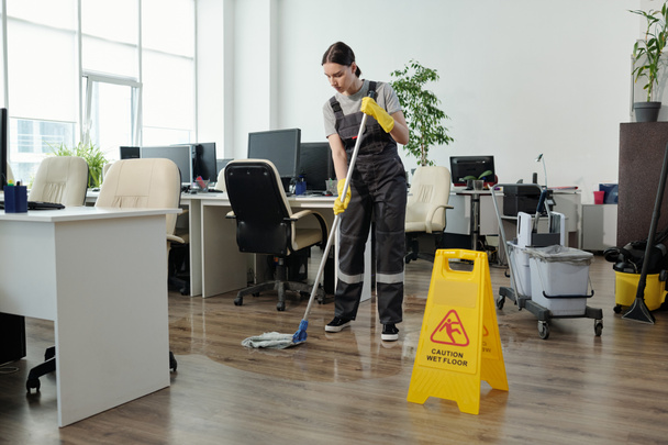 Young female cleaner in workwear using mop while cleaning floor in large modern openspace office with yellow plastic signboard in front - Photo, image