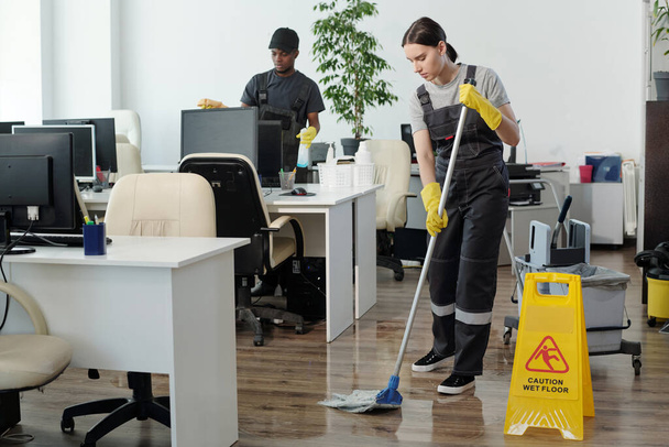 Young black man wiping computer monitors while Caucasian woman in coveralls and yellow gloves with mop cleaning floor in office - Foto, immagini