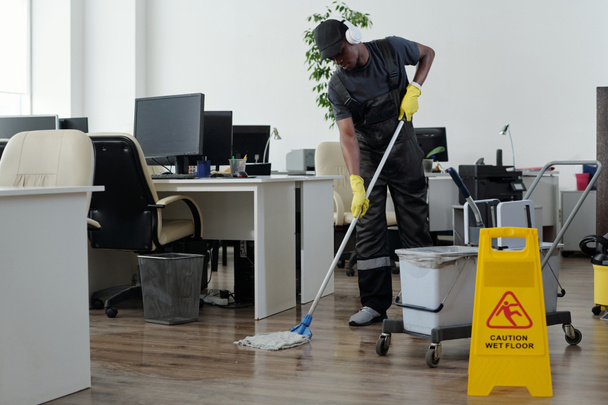 Joven hombre negro contemporáneo en el piso de limpieza de ropa de trabajo en la oficina de espacio abierto delante de letrero de plástico amarillo con precaución - Foto, imagen