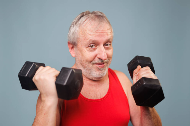 Ηλικιωμένος πολίτης στο γυμναστήριο αποδεικνύει ηλικία είναι μόνο ένας αριθμός Αυτή η εικόνα διαθέτει ένα αρσενικό συνταξιούχος ανύψωσης αλτήρες με αθλητική και ενεργητική στάση. Αλλά η κωμική έκφραση στο πρόσωπό του - Φωτογραφία, εικόνα
