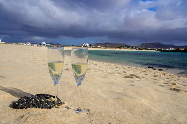 Δύο ποτήρια λευκό σαμπάνια ή cava αφρώδη οίνο σερβίρεται σε λευκή αμμώδη τροπική παραλία και μπλε ωκεανό, ρομαντικές διακοπές, το χειμώνα στον ήλιο Fuerteventura, Il Cotillo, Κανάρι, Ισπανία - Φωτογραφία, εικόνα