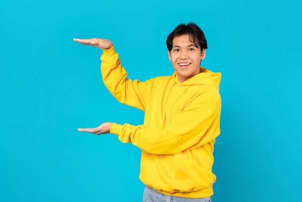 韓国の10代の男の子の手を笑顔サイズや広告を表示目に見えない製品を見てカメラの青のスタジオの背景に立って、黄色のパーカーを着て。このコンセプトを見てください - 写真・画像
