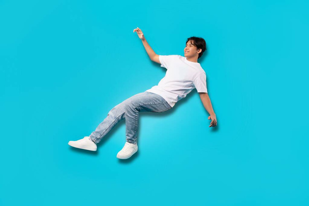 Happy Asian Teenager Boy Falling Posing In Mid Air Smiling Looking Aside Over Blue Background. Studio Shot Of Positive Teen Guy Experiencing Zero Gravity. Full Length - Foto, Bild