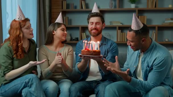 Etnikai sokszínű nők és férfiak party sapkában gratulálok férfi kaukázusi barátja bemutató meglepetés torta énekelni boldog születésnapot dal multiracionális többnemzetiségű emberek énekelnek ünnepi ünnepi ünnepség otthon - Felvétel, videó