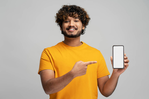Привлекательный улыбающийся индиец держит в руках мобильный телефон, делает покупки в интернете, указывая пальцем на пустой дисплей, изолированный на сером фоне. Современный счастливый азиатский хипстер, использующий мобильное приложение, заказывающий еду. Mockup - Фото, изображение