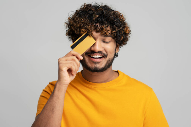 Sarı tişört giyen ve gri arka planda kredi kartı tutan gülümseyen çekici Hintli adamın portresi. Alışveriş, satış konsepti. - Fotoğraf, Görsel