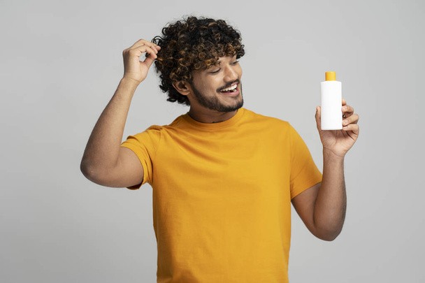 Glimlachende krullende Indiase man met fles met haarshampoo, op zoek naar cosmetische producten geïsoleerd op grijze achtergrond, studio schot. Haarverzorgingsconcept - Foto, afbeelding