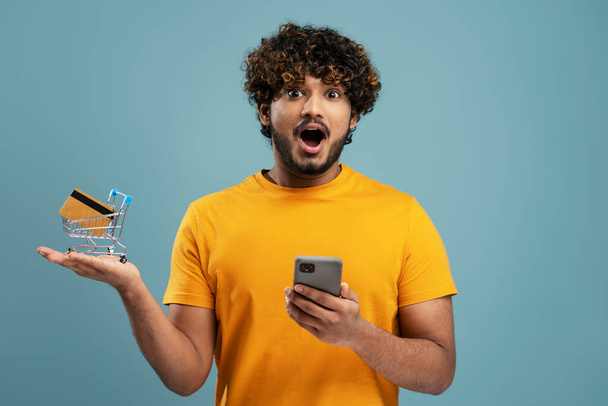 Fiatal izgatott indiai férfi mobiltelefonnal, kezében bevásárlókocsi hitelkártyával nézi kamera elszigetelt kék háttérrel. Vásárlás, értékesítés koncepciója - Fotó, kép