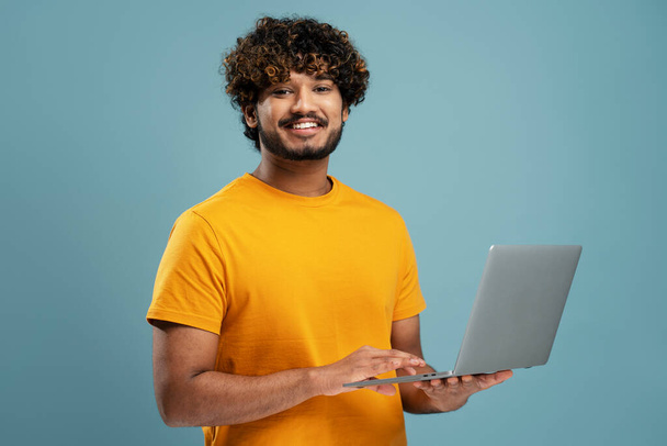 Smiling Indian man holding laptop computer working online looking at camera. Happy confident university student studying isolated on yellow background, online education. Portrait of happy programmer  - Photo, Image