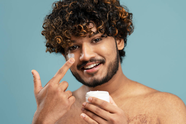 Close-up portret van aantrekkelijke metroseksuele Indiase man toe te passen anti-aging crème op zijn gezicht, kijken naar spiegel geïsoleerd op blauwe achtergrond. Huidverzorging, ochtend routine concept - Foto, afbeelding