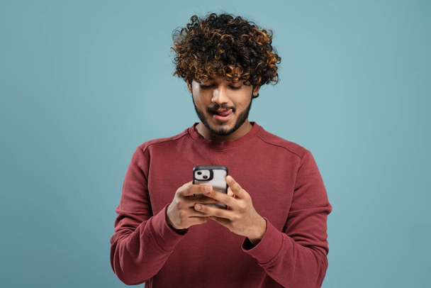 Hungry Indian man licking lips, holding smartphone using mobile app shopping online, ordering food on website isolated on grey background, copy space - Foto, Bild