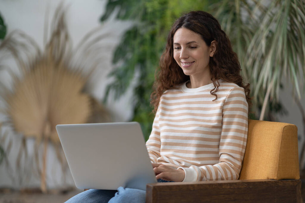 Radosna kobieta siedząca na fotelu relaksująca się przeglądając stronę internetową zakupów online. Happy girl surfing net, rozmowy z przyjaciółmi podczas wolnego czasu w kawiarni. Młoda kobieta wykonująca pracę badawczą dla biznesu. - Zdjęcie, obraz