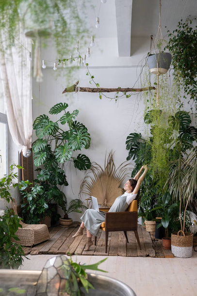 Happy relaxed woman remote worker resting, stretching arms while working at nature-inspired home office filled with many different exotic indoor plants, sitting with laptop on chair in home garden - Φωτογραφία, εικόνα