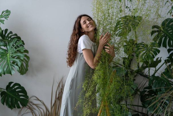 Mutlu bir kadın bahar gününü yaprak bitkisine sarılarak, gülümseyerek ve evinin bahçesinde merdiven üzerinde durarak geçiriyor. Kapalı alandaki saksı çiçekleri ve ev ormanı. Stres giderici olarak bitki bakımı. - Fotoğraf, Görsel