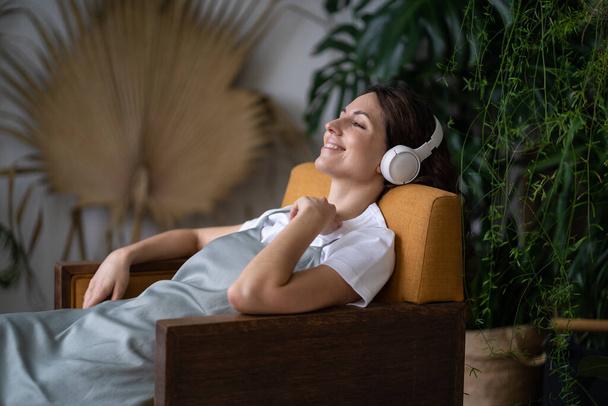 Relaxed girl lies surrounded by exotic green plants in the city botanical garden in wireless headphones. Smiling woman resting after working day listening to music, interesting podcasts in earphones. - Foto, Bild